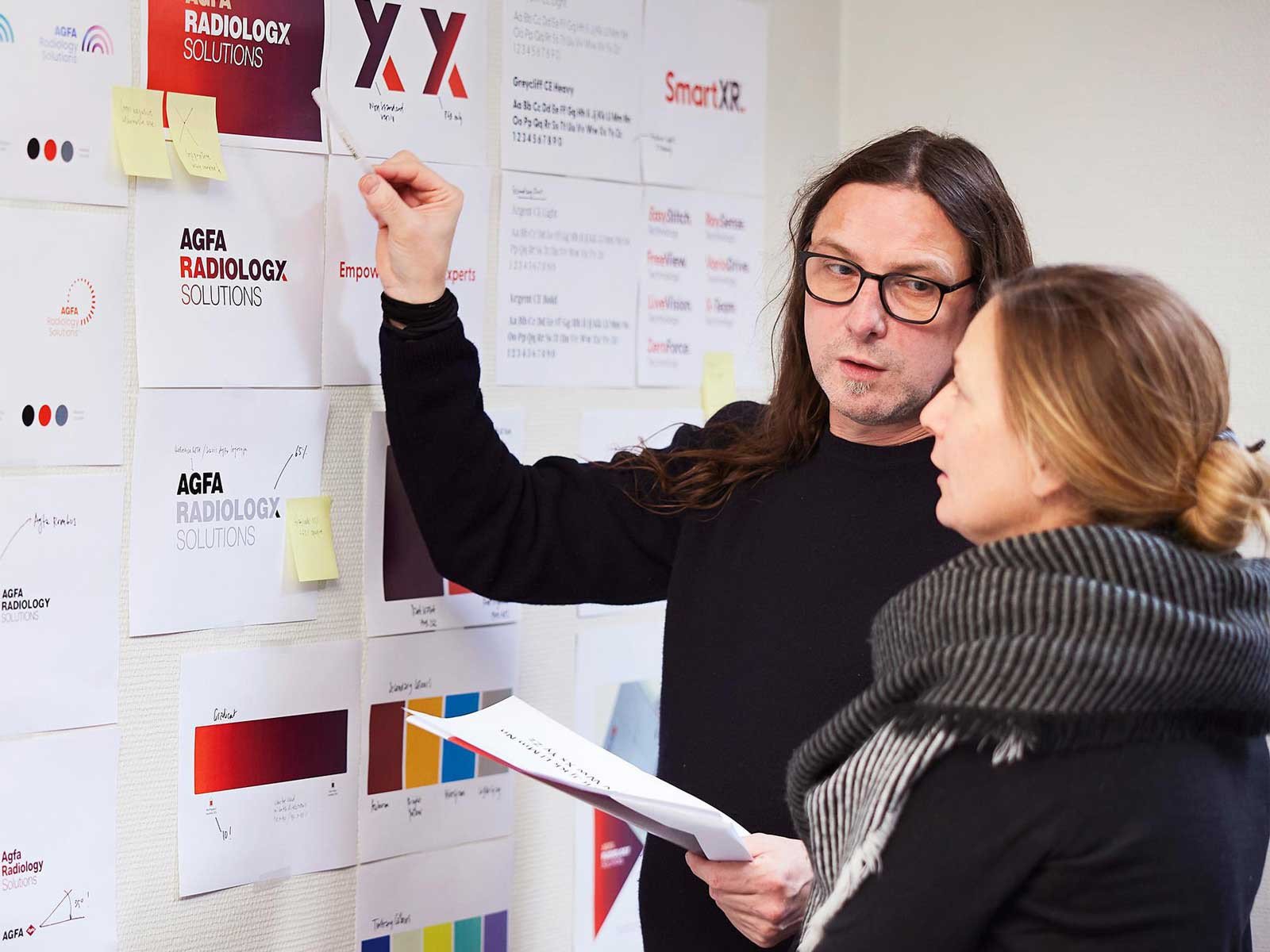 Twee personen bespreken branding concepten voor Agfa Radiology Solutions aan een muur vol logo-ontwerpen, kleurstalen en notities.