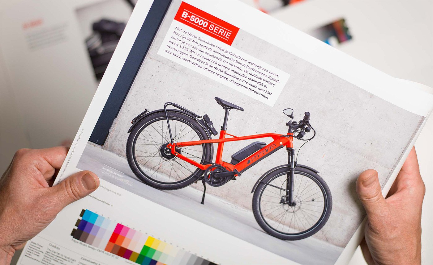 Brandle - Beeldbewerking na fotografie voor catalogus van Norta Handen die een drukproef van een Norta-catalogus vasthouden, met een foto van een feloranje elektrische fiets en kleurstalen onderaan het drukvel.