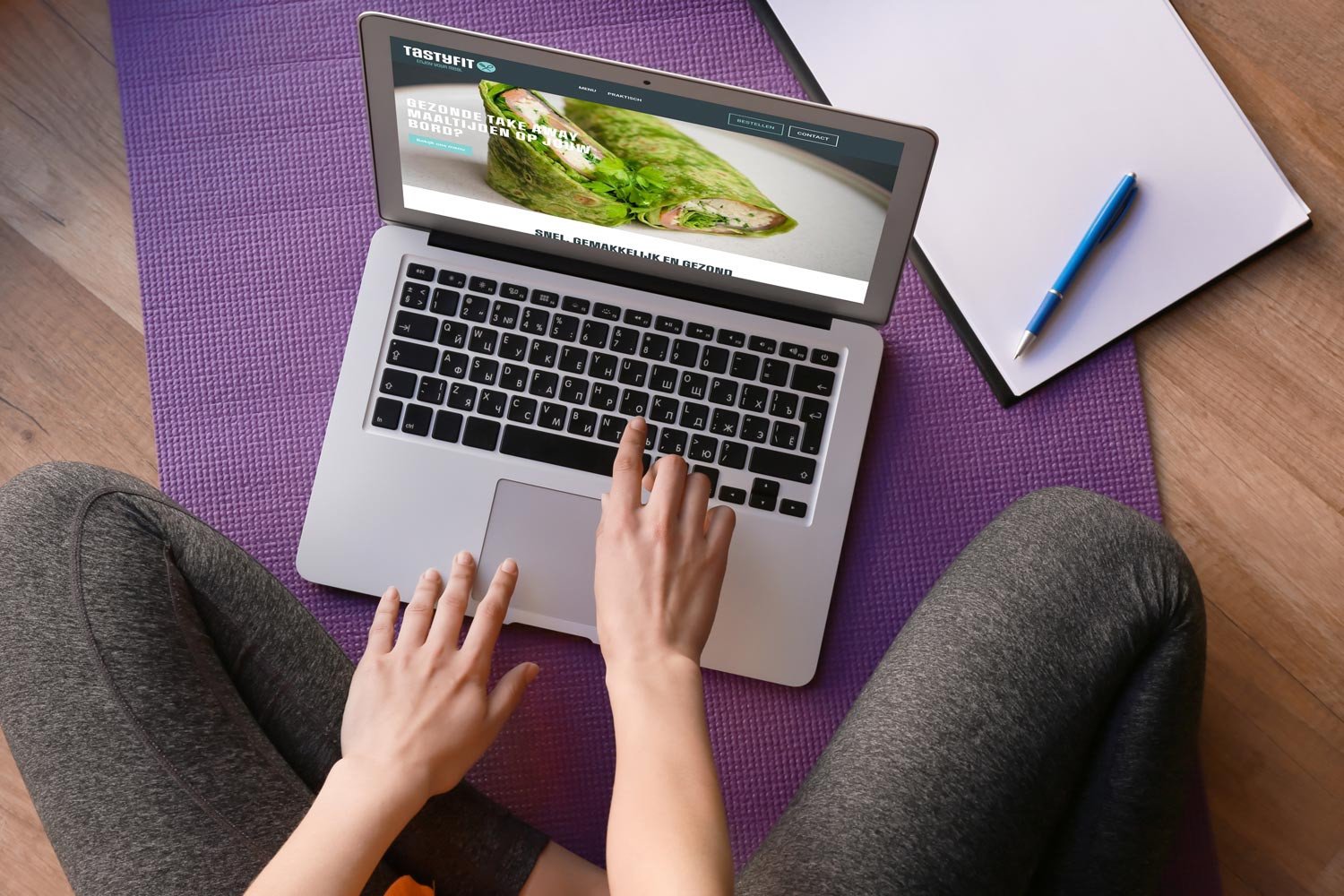 Persoon werkt op laptop met Tastyfit-website, zittend op yogamat met notitieboek en pen ernaast.