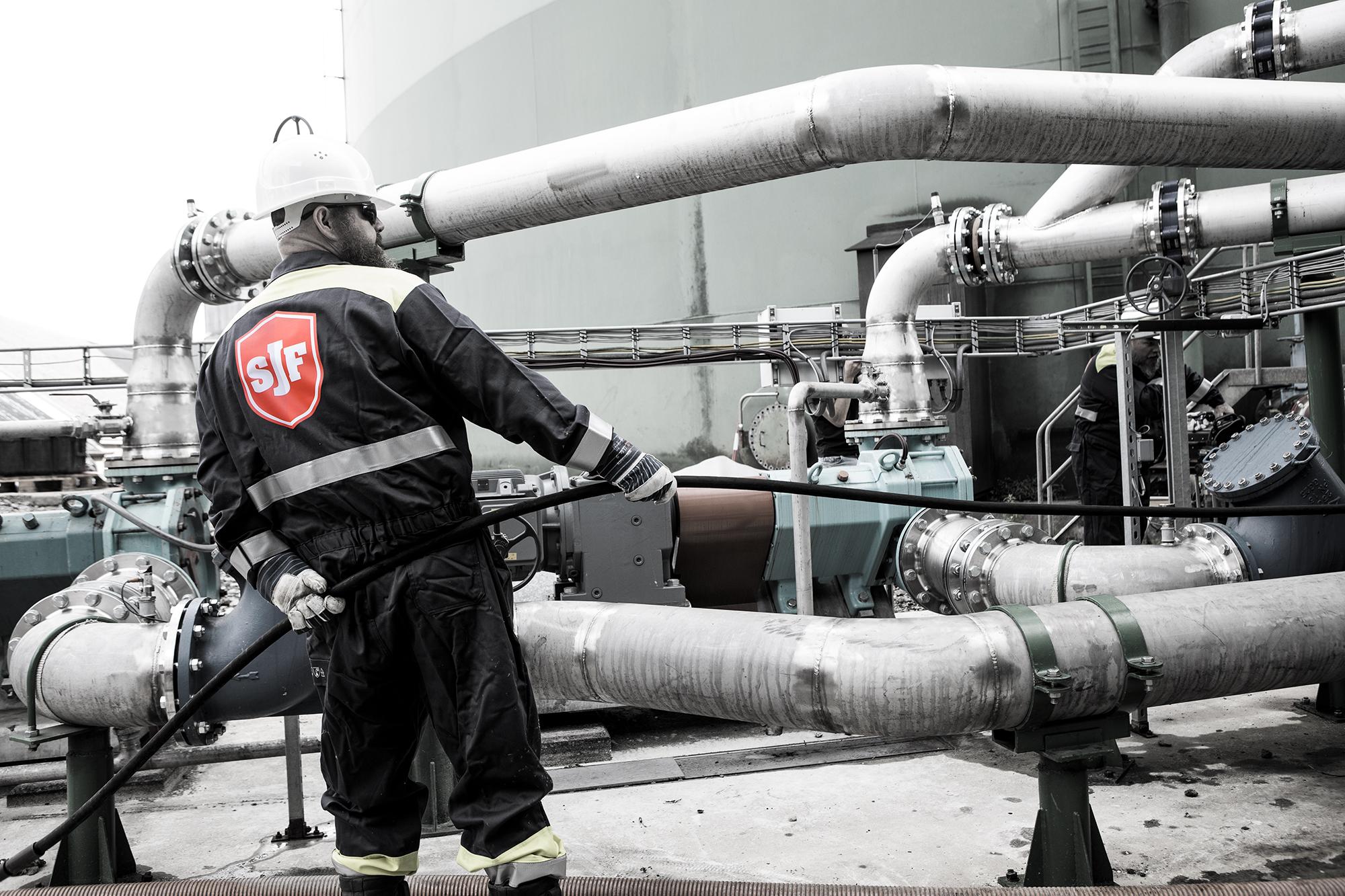 Medewerker van SJF in beschermende werkkleding werkt aan industriële leidingen en installaties in een technische omgeving.